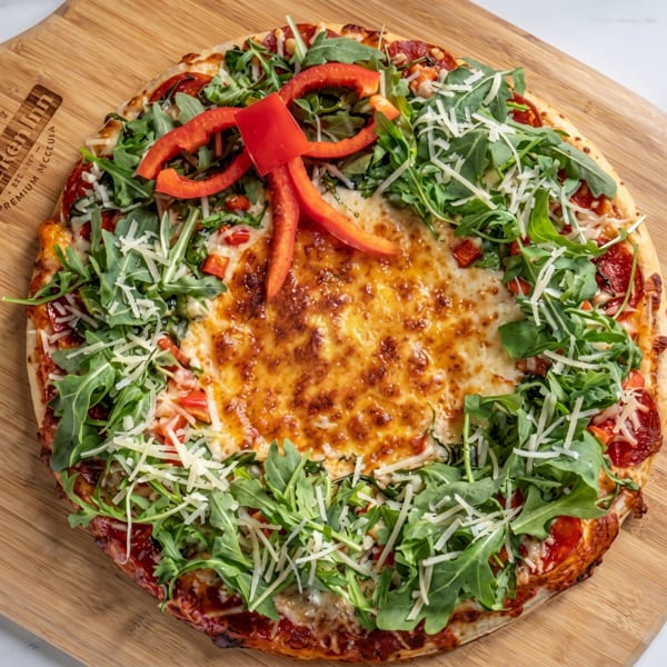 A pizza with a ring of arugula, shredded cheese, and red bell pepper slices on a wooden board.